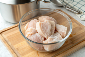 A top-down, overhead view of a clear glass bowl filled with several rock-solid, icy frozen chicken breasts. The bowl sits on a wooden cutting board with a stainless-steel pot in the background. The lighting is bright and natural, emphasizing the texture of the frozen chicken